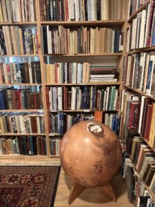 A wooden bookshelf of at least five levels occupies two corners of a room. A dark red patterned carpet is on the floor in the left foreground. A large brown globe stands to its right.