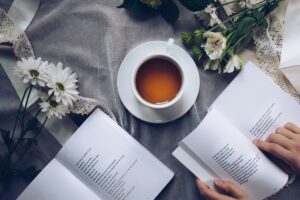 Two open books, one with human hands touching it, on a table. Flowers and a mug with tea are also on the table.