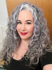 Headshot of a person with long, curly silver hair; bright red lipstick, and wearing a black T-shirt.